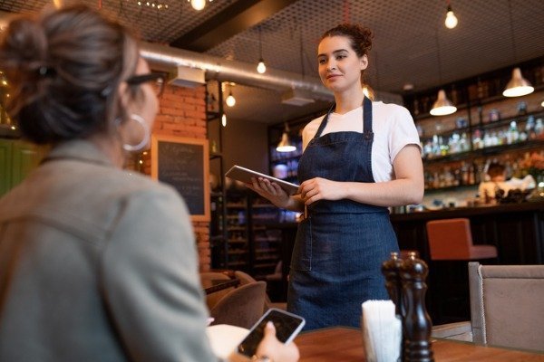 waitress-wearing-an-e-holster-server-apron-holding-a-tablet-while-standing-by-the-table-in-a-restaurant